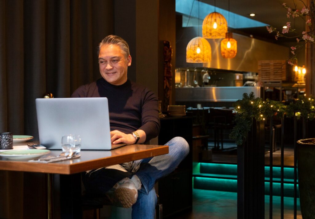 Een man zit aan een tafel in een gezellige bar, met een laptop voor zich, terwijl hij lacht en ontspannen kijkt.