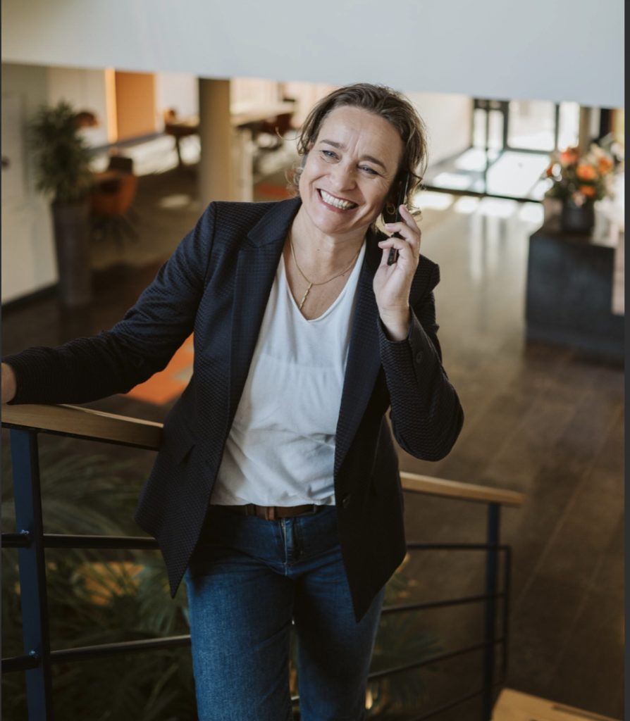 Vrouw in zakelijke kleding leunt lachend op een trapleuning terwijl ze belt met haar mobiel in een modern kantoorpand. Deze afbeelding ondersteunt het thema zakelijk mobiel werken in een professionele omgeving.